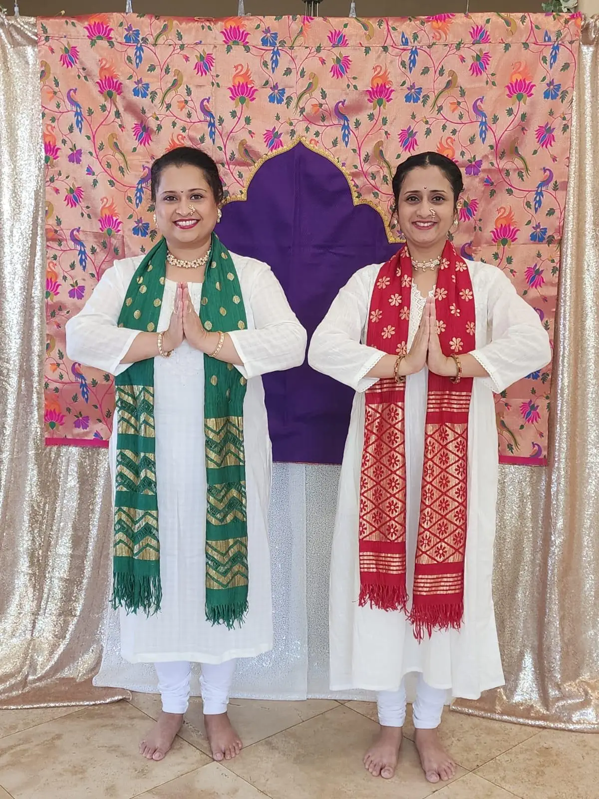 Manaswini Arts co-founders Ashwini Katre-Kulkarni and Manali Phadke in white kurtas with green and red dupattas, greeting the audience with a namaste before a Paithani-patterned backdrop.